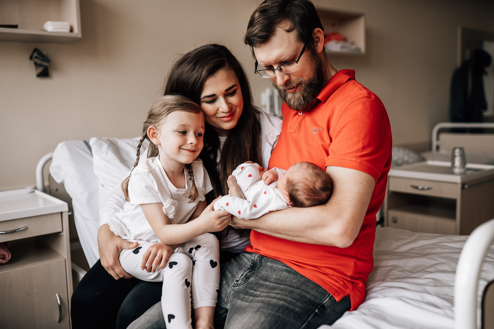 Fotografie z porodnice. Tatínek drží v náručí miminko. On, maminka i starší dcera sedí společně na nemocničním lůžku a radují se z novorozeného miminka.